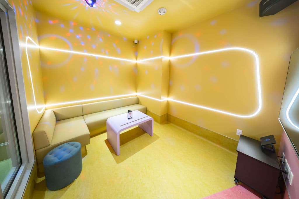 Small karaoke room with a yellow corner sofa, teal stool, and glowing LED light strips