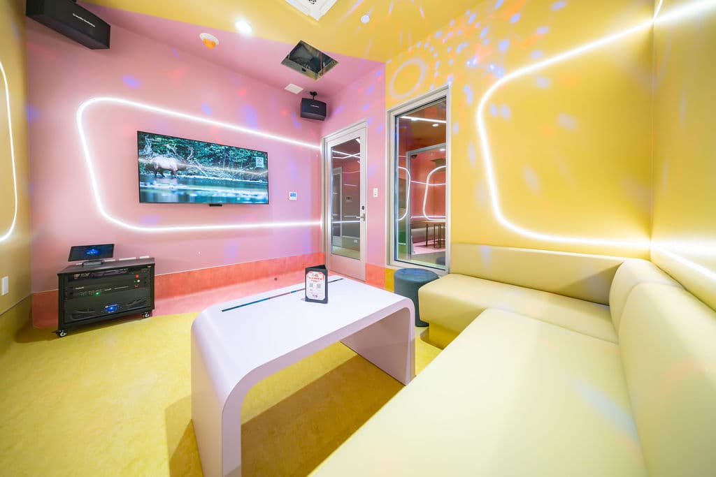 Small karaoke room with a pink screen wall, glass door, and yellow corner sofa