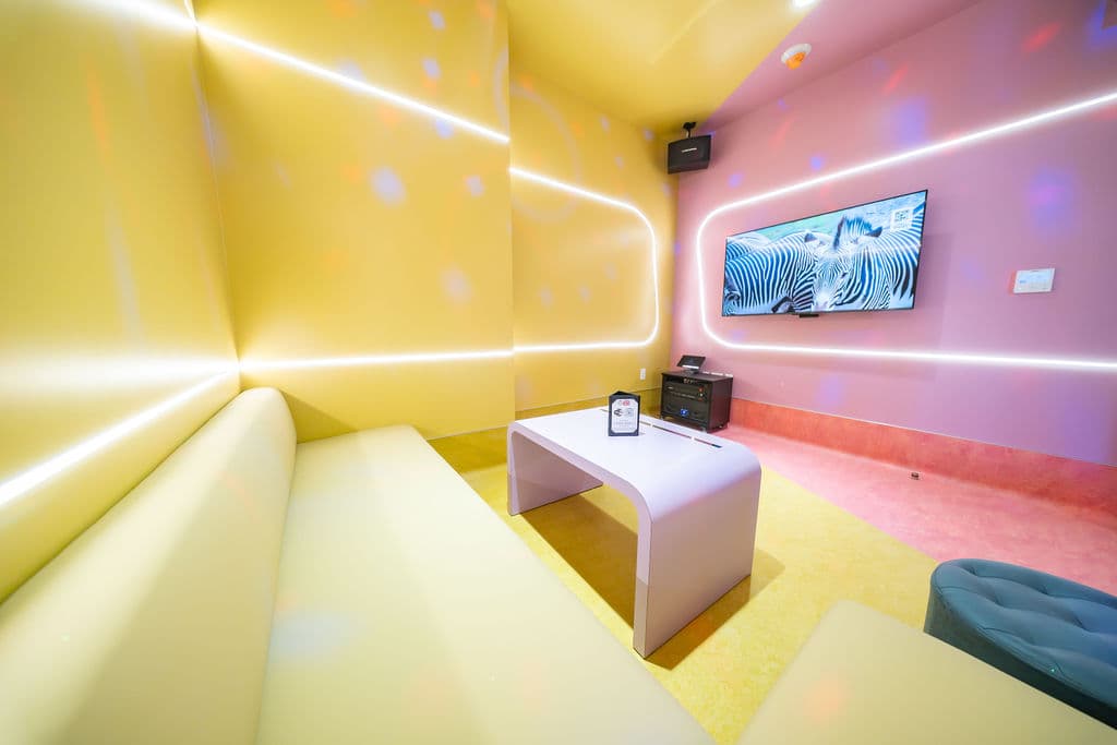 Small karaoke room with a yellow sofa facing a wall-mounted screen on a pink wall