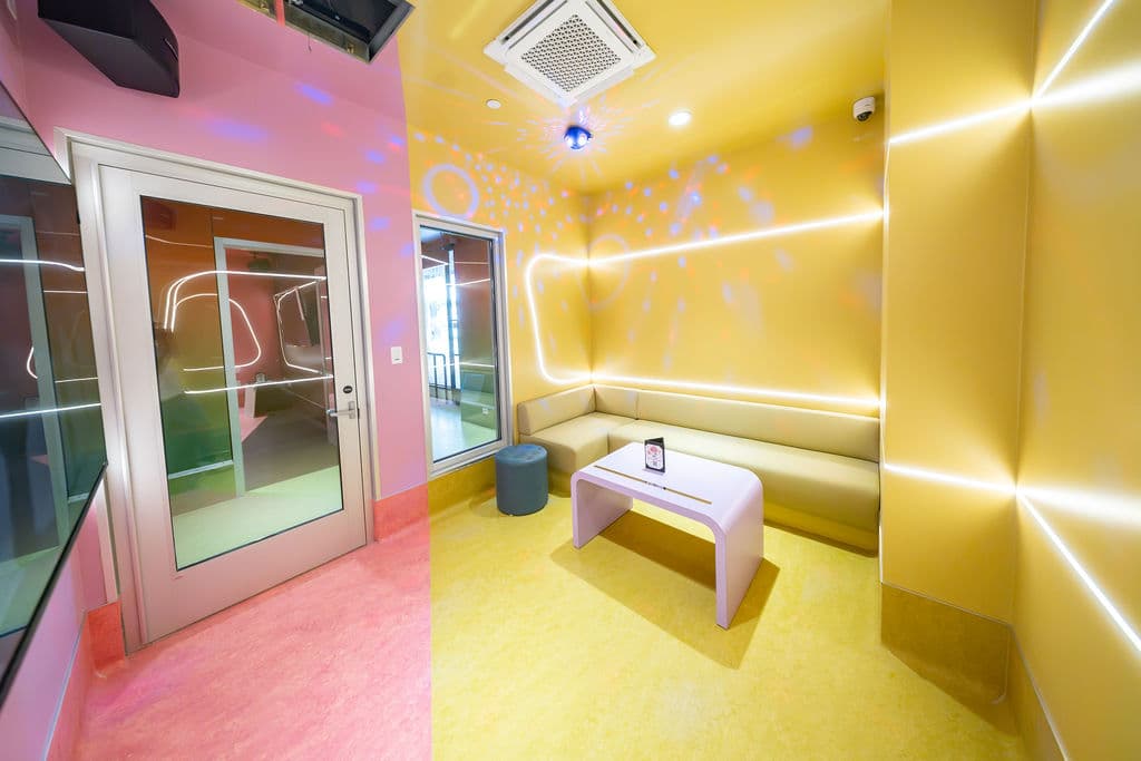 Small karaoke room seen from the pink entry door, with a yellow sofa and white table