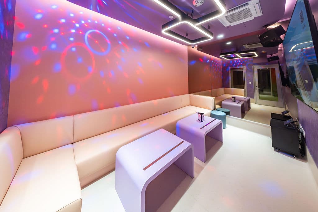 Large karaoke room with a cream sofa and lavender tables under disco lights